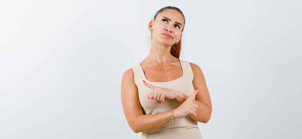 young girl pointing opposite directions with index fingers in beige top, black pants and looking pensive , front view.