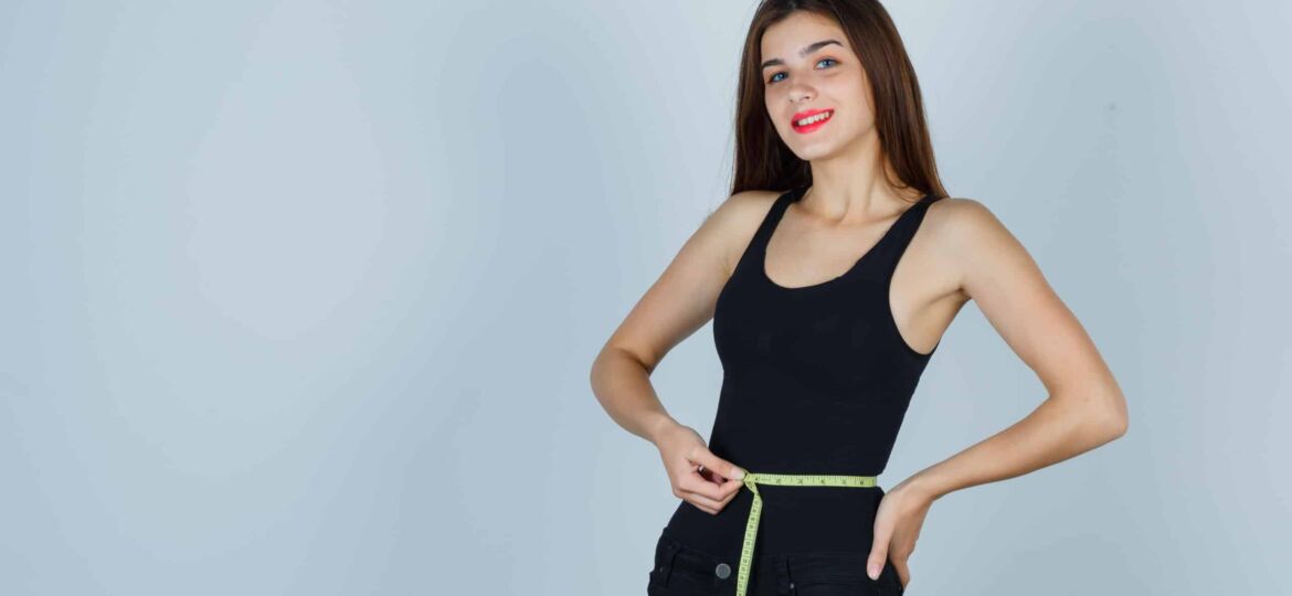 young girl holding measuring tape around her waist in black top, pants and looking confident. front view.