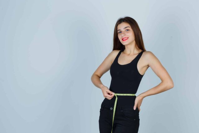 young girl holding measuring tape around her waist in black top, pants and looking confident. front view.