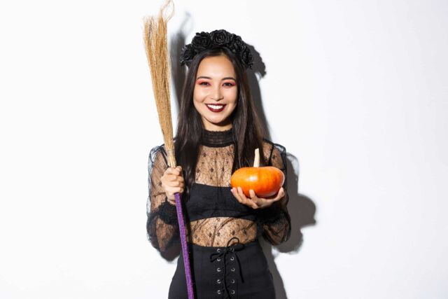 Image of beautiful asian woman dressed-up as a witch for halloween party, holding broom and pumpkin, standing over white background.