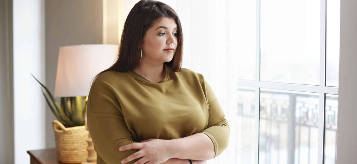 People, lifestyle and body positivity concept. Beautiful young brunette female with extra pounds standing by window at home, crossing arms on her chest, having thoughtful pensive look, pondering