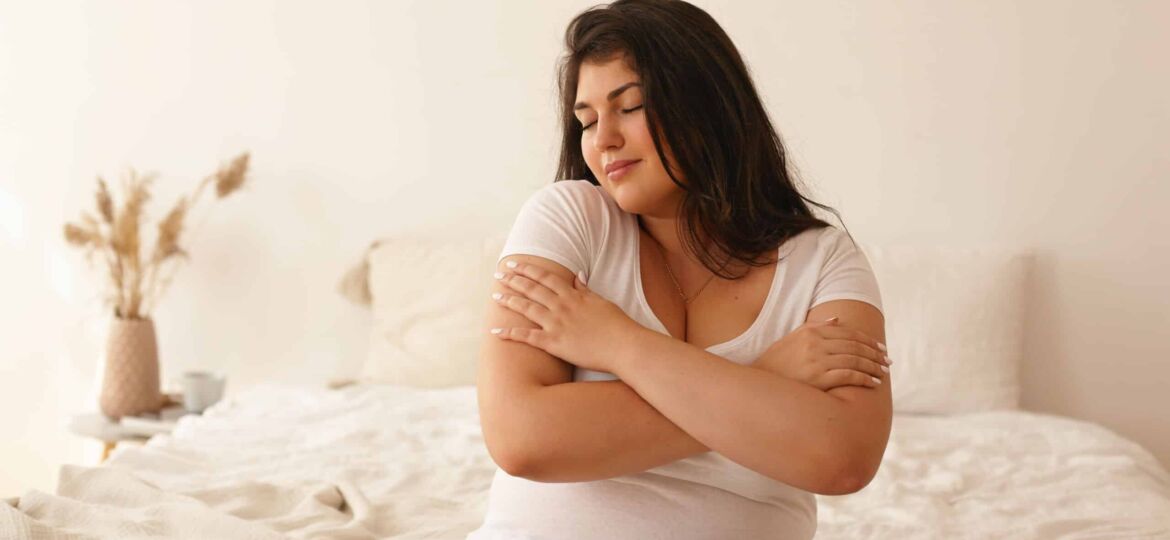 Indoor shot of gorgeous overweight young Latin woman in casual clothes sitting in bedroom on white bed, closing eyes, smiling, touching her soft skin after applying moisturizing body cream