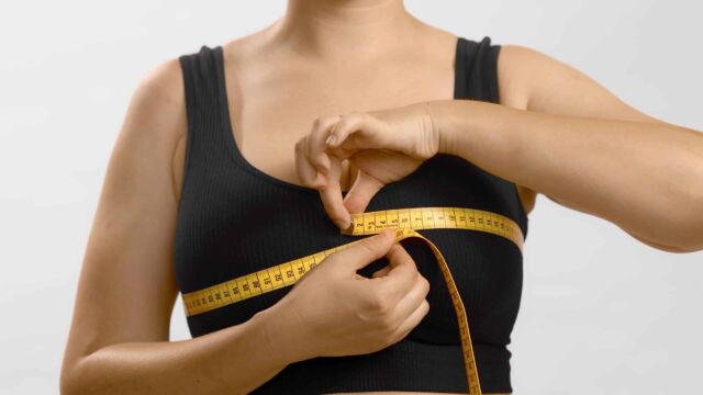 Caucasian young woman in black sport bra measuring her breast with a tape. Studio high quality photo on white background.