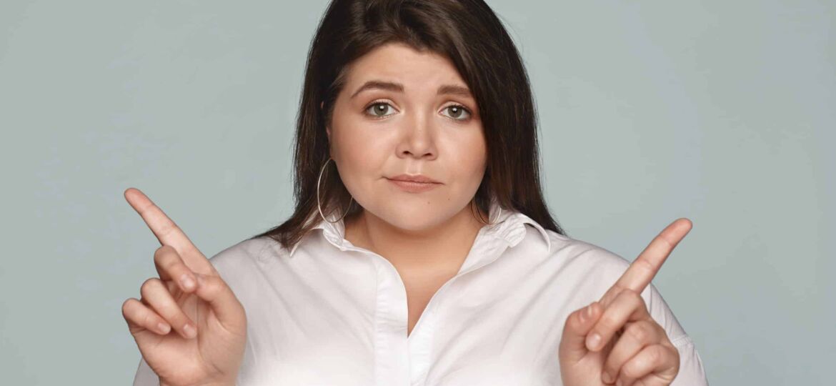 Puzzled young dark haired businesswoman with chubby cheeks and curvy body pointing her index fingers at opposite directions, can't choose between two business concepts, having doubtful look