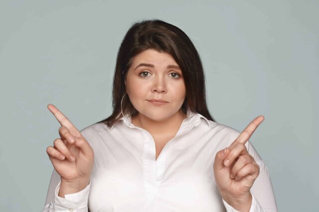Puzzled young dark haired businesswoman with chubby cheeks and curvy body pointing her index fingers at opposite directions, can't choose between two business concepts, having doubtful look