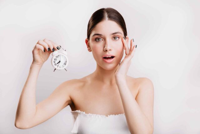 Adult woman with elastic healthy skin looking at camera, holding small white alarm clock.