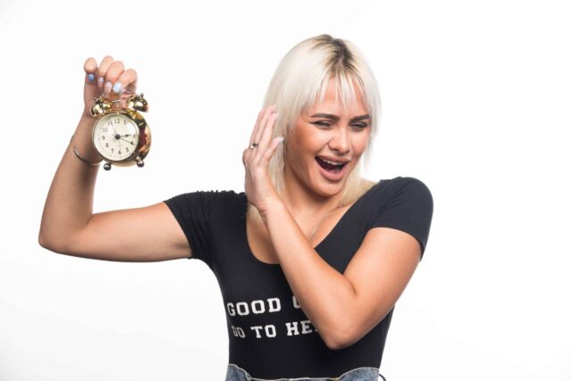 Young woman holding clock while pointing her hand towards it on white background