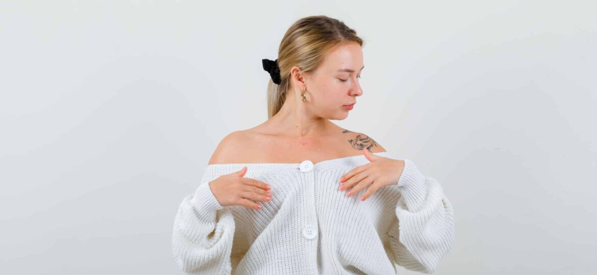 Portrait of young blonde woman posing while holding hands over chest in cardigan, jeans and looking graceful front view