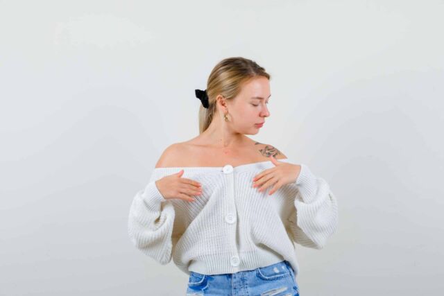 Portrait of young blonde woman posing while holding hands over chest in cardigan, jeans and looking graceful front view