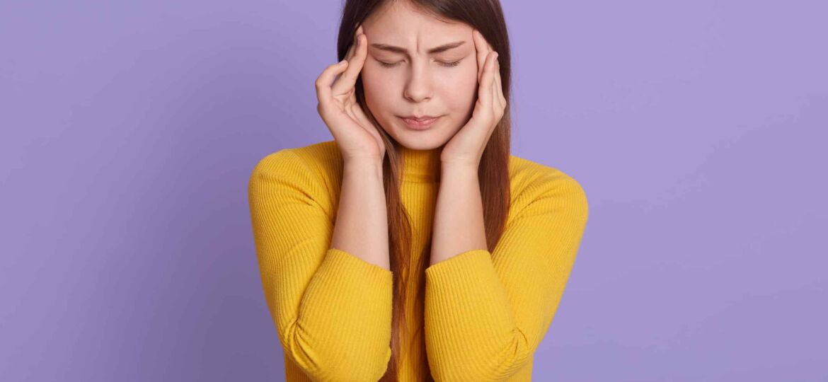 Portrait of beautiful young woman keeping hands on temples, suffering from terrible headache, keeping eyes closed, looks sad, feels unwell, wearing yellow casual shirt.