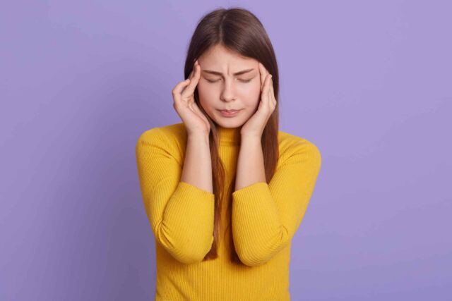 Portrait of beautiful young woman keeping hands on temples, suffering from terrible headache, keeping eyes closed, looks sad, feels unwell, wearing yellow casual shirt.