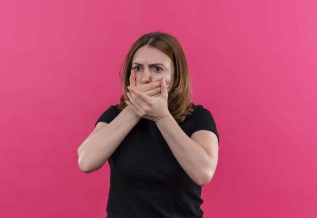 anxious young casual woman with hands on mouth on isolated pink background with copy space