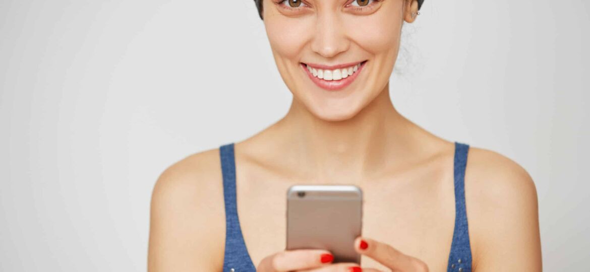 Beautiful smiling casual woman holding smartphone, looking and smiling at the camera isolated against white studio wall background with copy space for your text or advertising content. Film effect